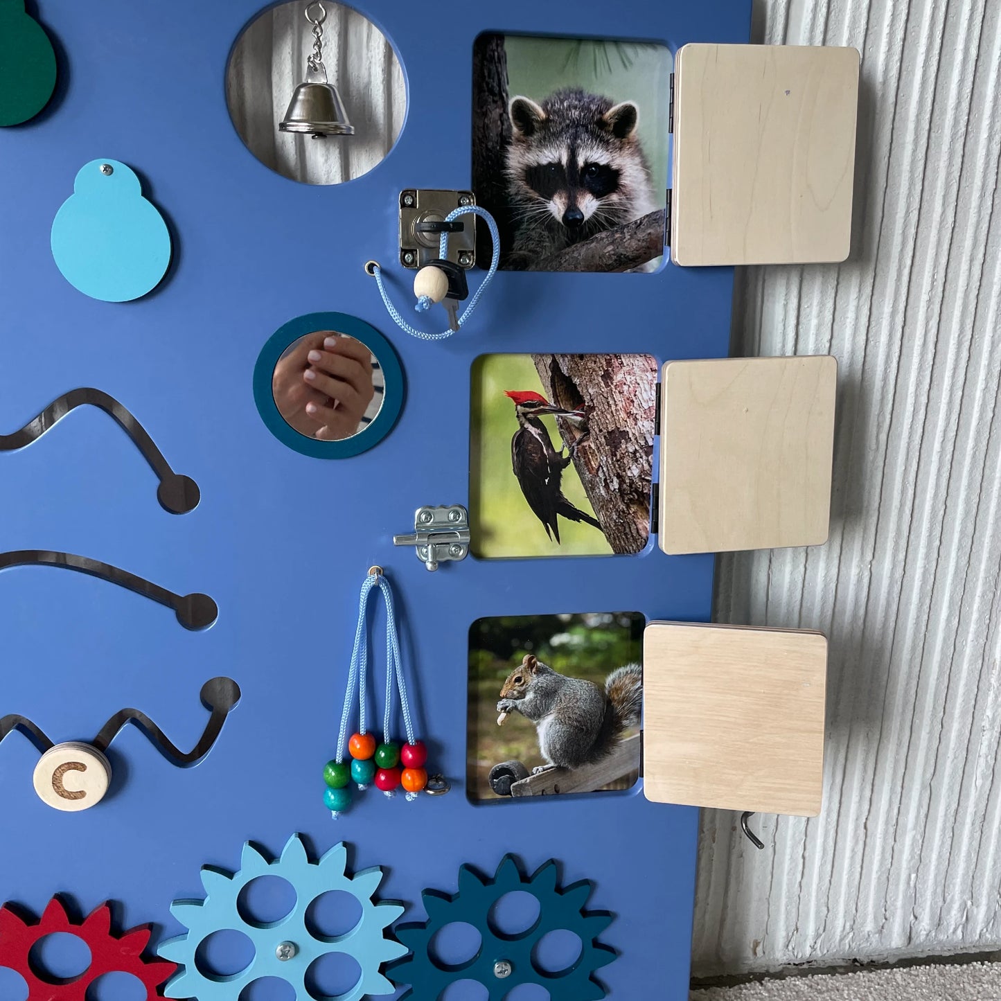 Close-up of sensory wall panel with flip-up animal picture flaps, small mirror, bell, colorful beads on cords, and red and blue gears for interactive sensory play