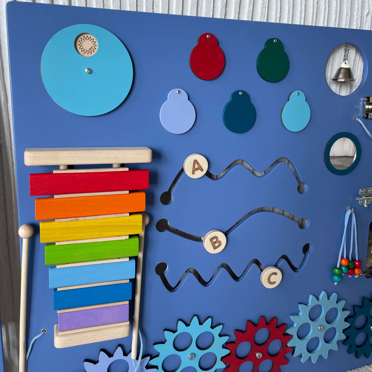 Close-up of sensory wall panel featuring rainbow xylophone, tracing paths with letters A-B-C, color matching elements, mirror, bell, and colorful gears for fine motor skill development