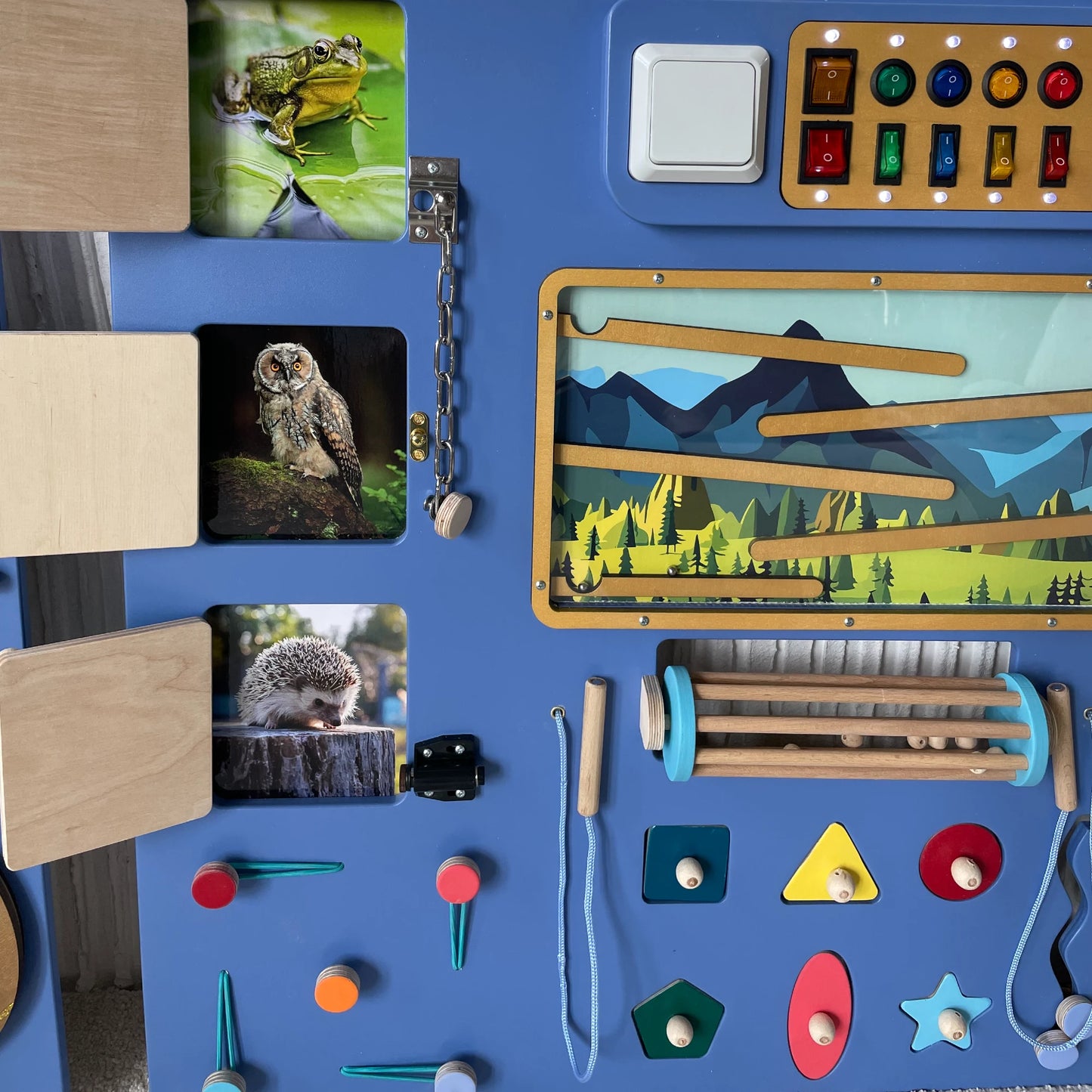 Close-up of wooden sensory wall showing animal cards behind doors, mountain-themed marble maze, toggle switches, and fine motor skill activities
