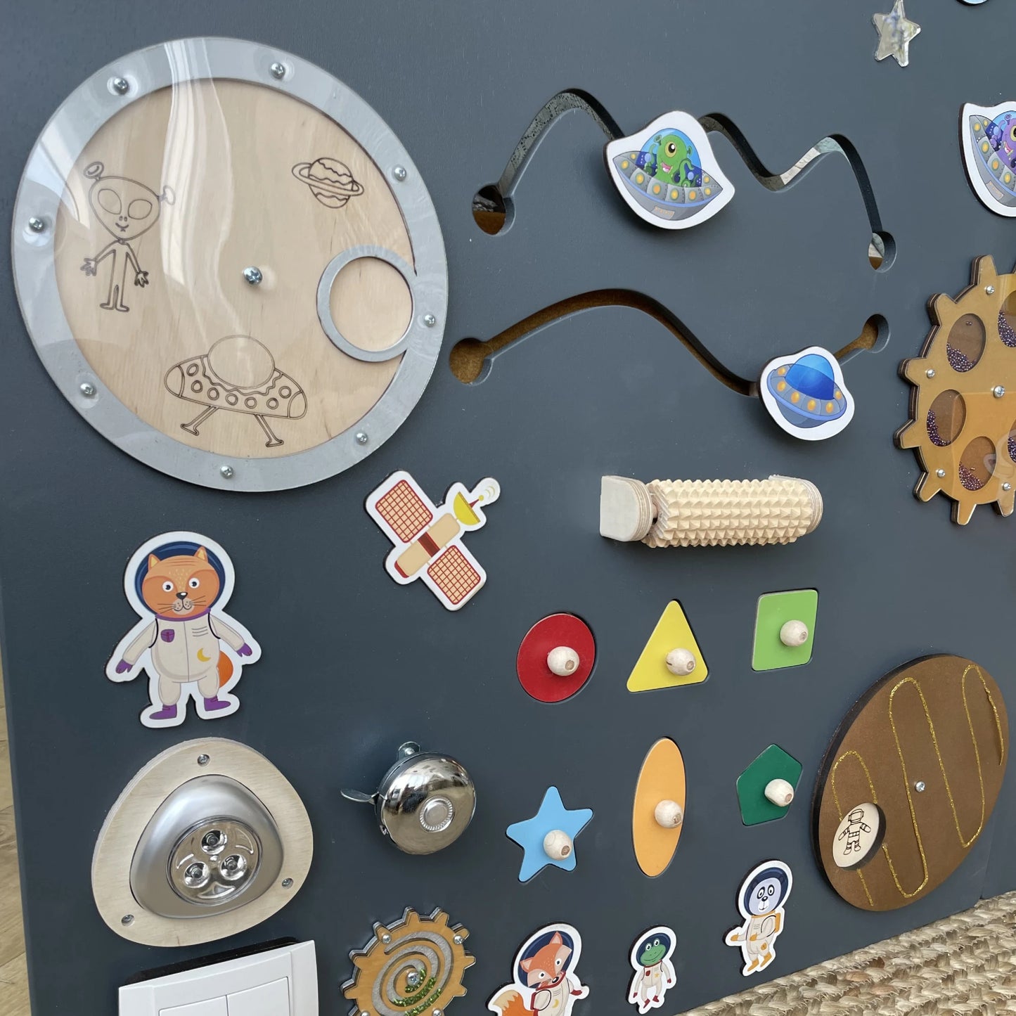 Space-themed sensory wall panel with UFO sliders, tactile textures, geometric shape puzzles, bell, and alien illustrations for fine motor skills and visual stimulation