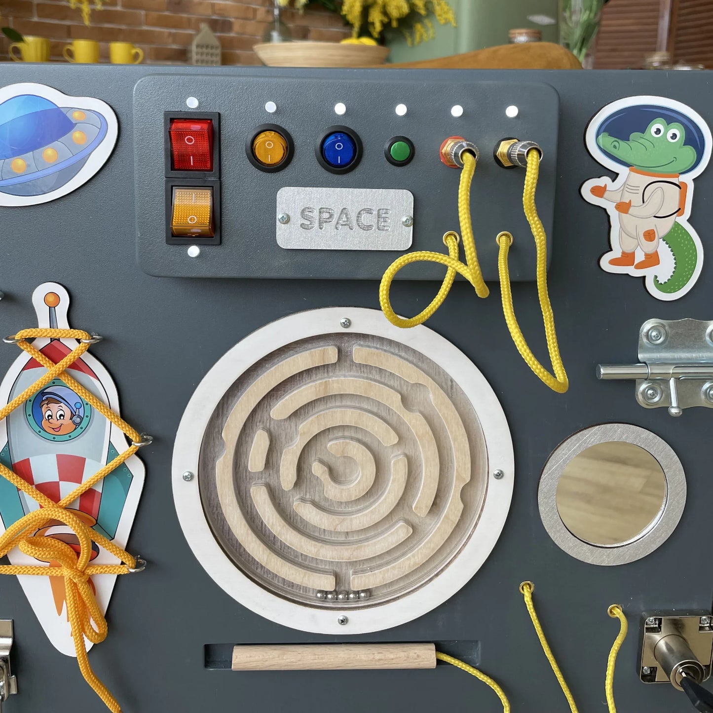 Close-up of a space-themed sensory wall panel showing LED switches, wire maze, space-themed stickers, and tactile fine motor activities on a dark grey background