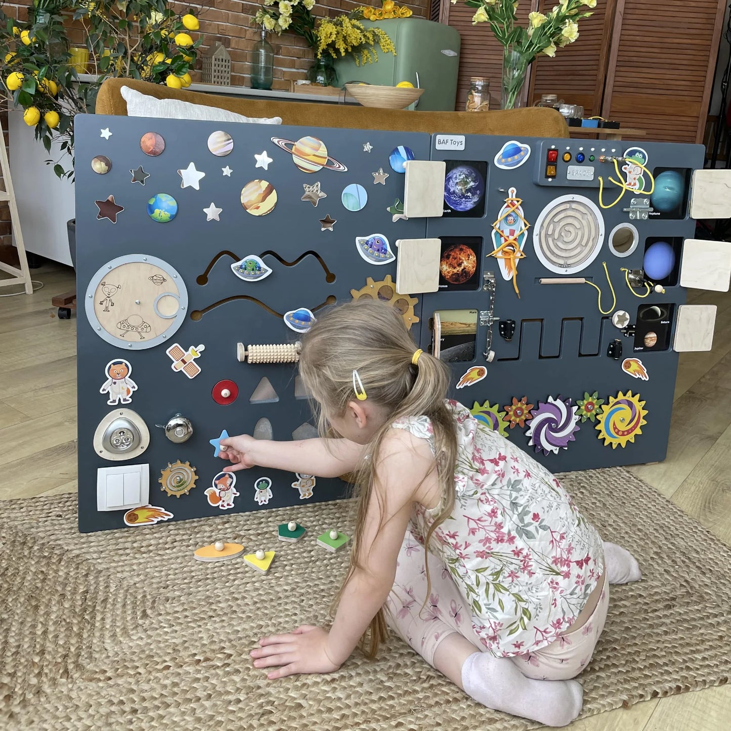 Little girl playing with space-themed sensory wall panel featuring planets, shape puzzles, gears, and tactile elements designed for cognitive and motor skill development