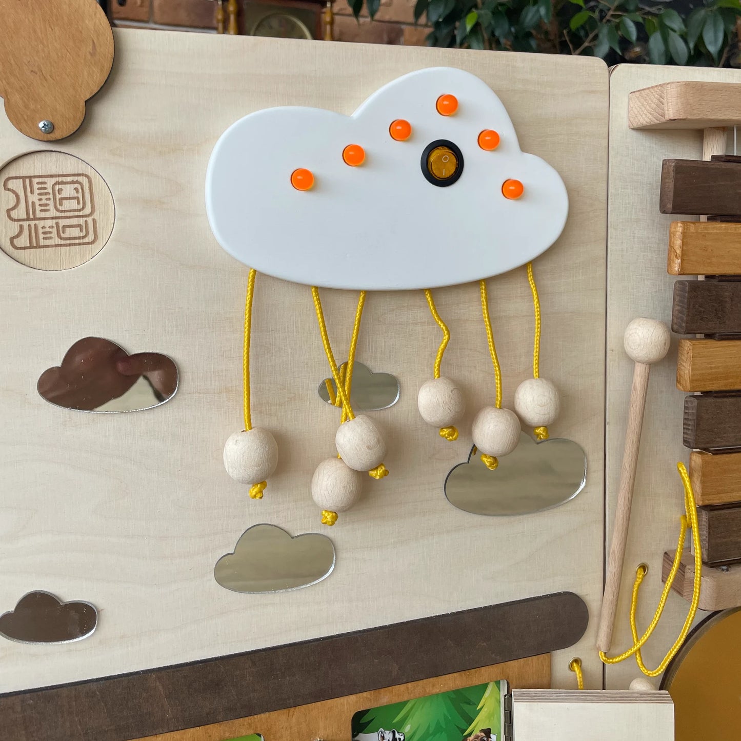 Close-up of a white LED cloud element with wooden raindrop beads and yellow strings on a train-themed sensory wall panel for kids