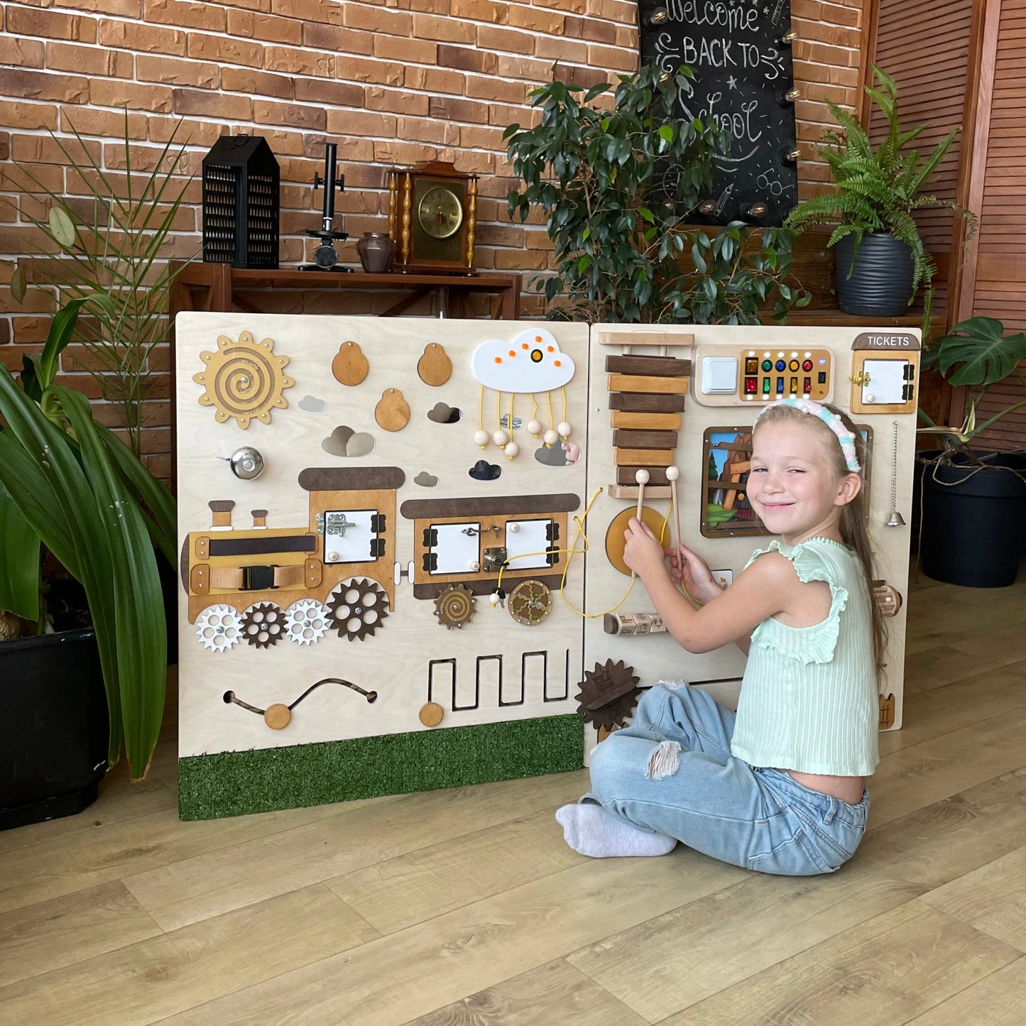 Smiling girl plays with train-themed sensory wall panel featuring wooden xylophone, gears and fine motor elements. Ideal for preschool, special needs therapy or classroom learning spaces