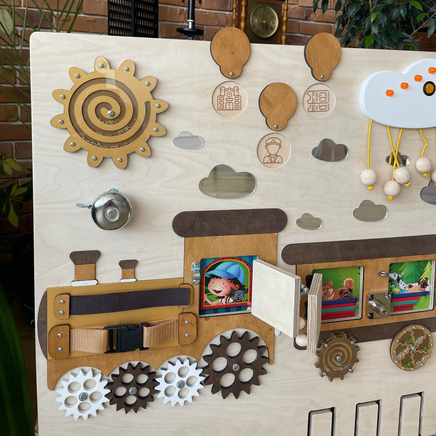 Close-up of the left section of a train-themed sensory wall panel with sun spiral, bell, belt fastener, gears, and interactive photo windows for kids