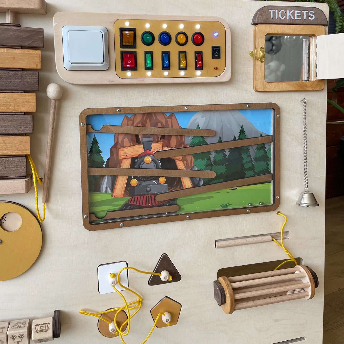 Close-up of the right part of a train-themed sensory wall panel featuring a switchboard with lights, ticket window, maze with train illustration, bell, and lacing activities