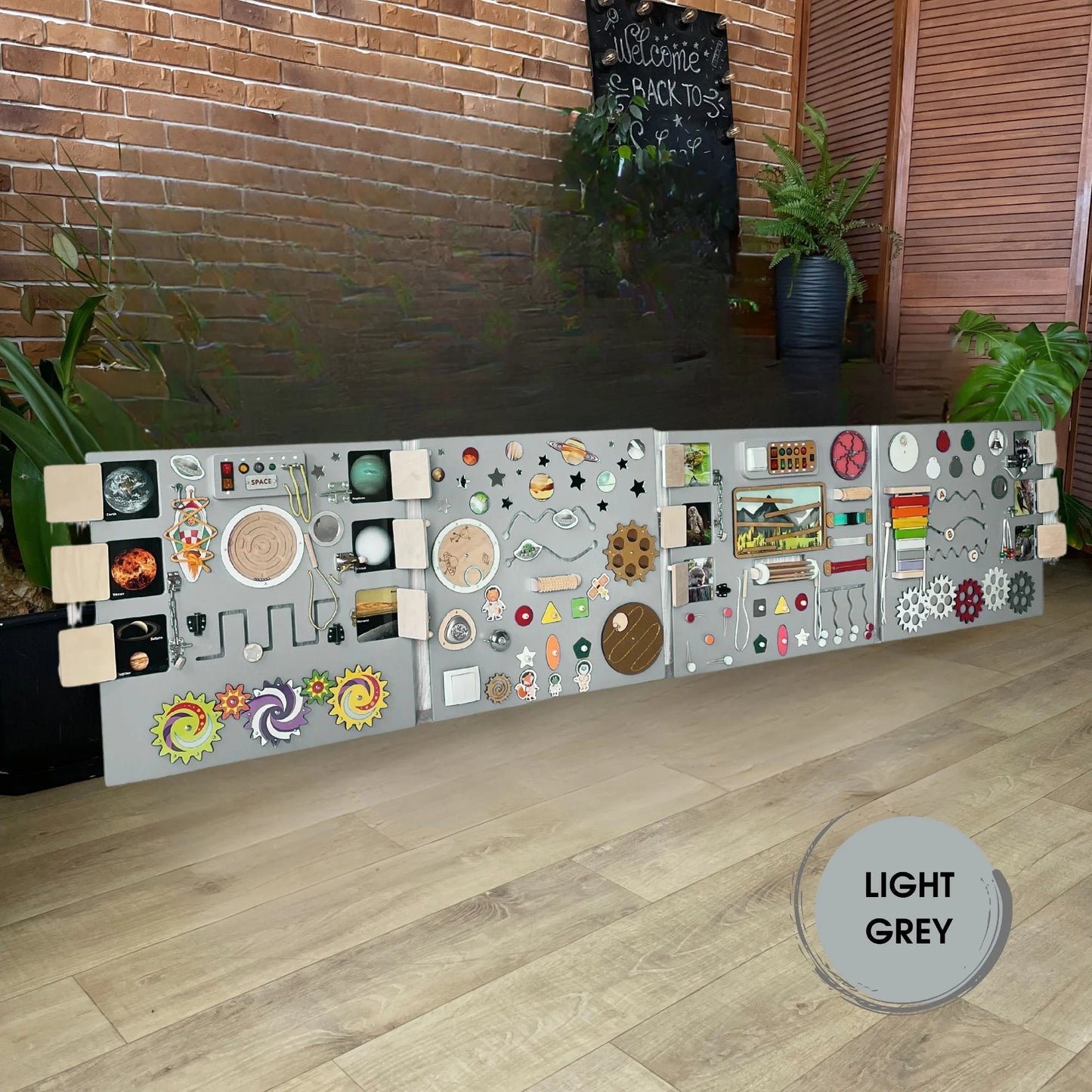 Light grey XXL sensory wall panel with 4 sections, LED lights, and 75 activity elements for kids and adults, displayed against a brick wall background