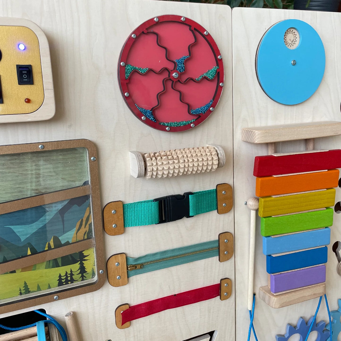 Close-up of buckles, zipper, spinning glitter disk, and textured roller on wooden sensory board.