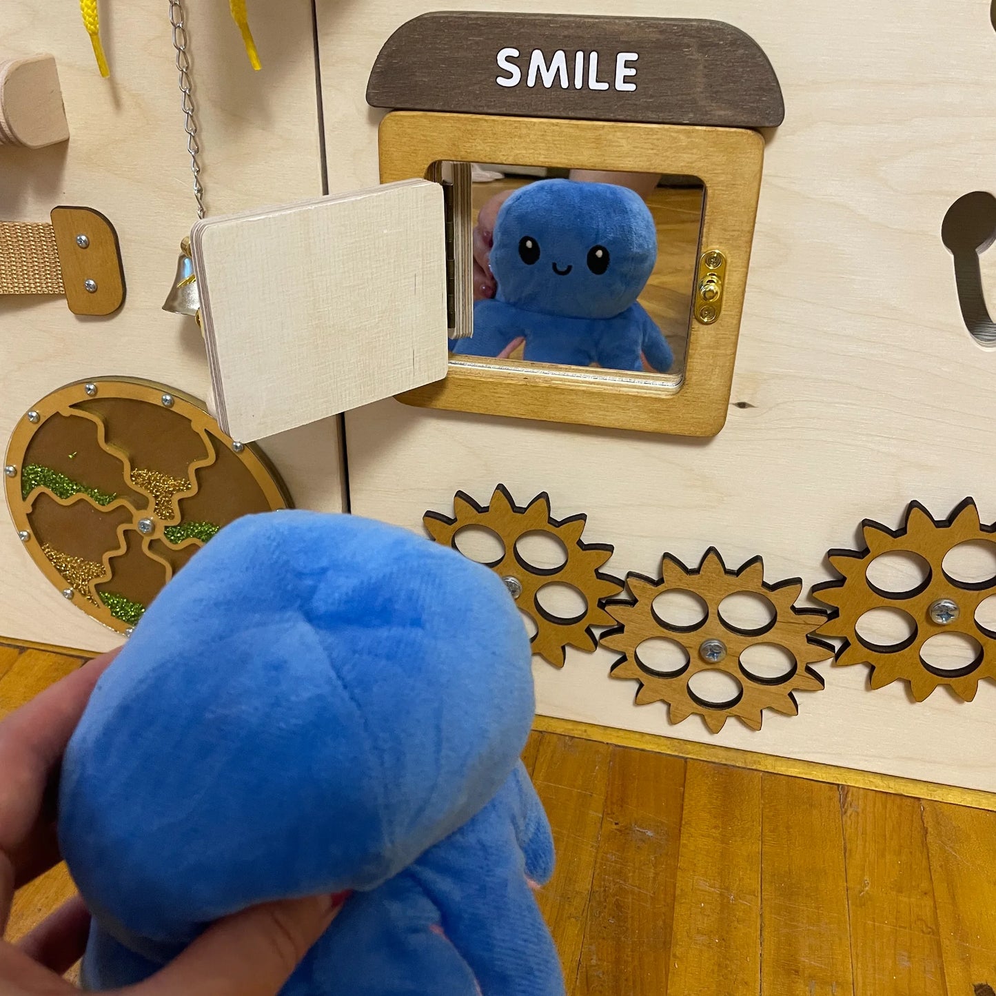 Plush octopus toy reflected in sensory mirror on busy board with gears and smiley face detail
