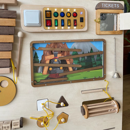 Close-up of the right part of a train-themed sensory wall panel featuring a switchboard with lights, ticket window, maze with train illustration, bell, and lacing activities
